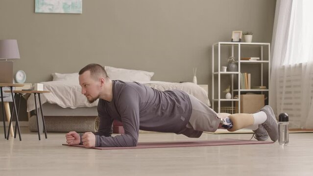 Slowmo of concentrated young Caucasian man with prosthetic leg doing plank exercise on training mat during home workout