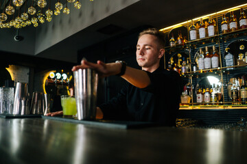 Low-angle view of young barman in black clothes making refreshing alcoholic cocktail standing behind bar counter in modern dark nightclub, on background of shelves with different alcoholic drinks