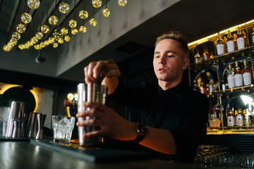 Close-up low-angle view of focused bartender making refreshing alcoholic cocktail standing behind bar counter in modern dark nightclub, on background of shelves with different alcoholic drinks