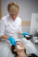 Doctor cosmetologist or dermatologist making face mask in cosmetology salon. Professional Beautician applying face mask on caucasian woman face lying on bed in bathrobe.