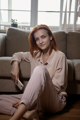 Fototapeta premium Caucasian young good looking woman with red hair sitting on the floor near the sofa in room and tapping or playing on the smartphone.