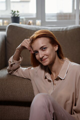 Caucasian young good looking woman with red hair sitting on the floor near the sofa in room and Woman looking at camera, smiling and laughing. Joy, warmth, happiness.