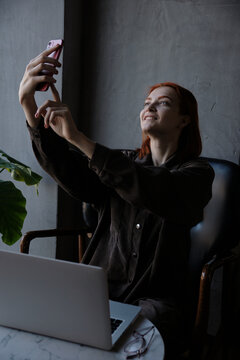 A red-haired woman takes pictures of herself on the phone while sitting in a cafe.