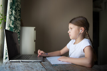 Fototapeta premium Cute european kid girl with two pigtails studying at home at the table with a laptop and headphones, distance learning and tutor