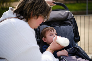 Mother giving a bottle of milk to her son while enjoying the day in the park.