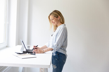 pretty young blonde business woman stands by her ergonomic height adjustable work desk and works on...