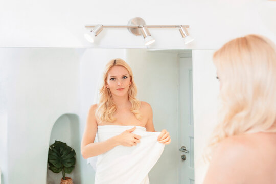 blond woman in a white bathroom looking at her reflection in the mirror, wearing a towel