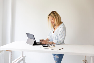 pretty young blonde business woman stands by her ergonomic height adjustable work desk and works on...