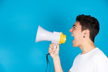 Hispanic teenager boy shouting on megaphone or speaker to copy space isolated on blue background.