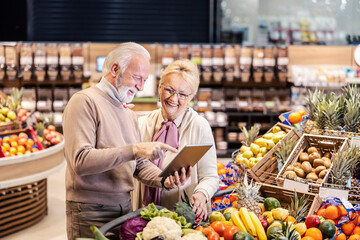 A happy senior couple using tablet for groceries list and shopping at the supermarket.