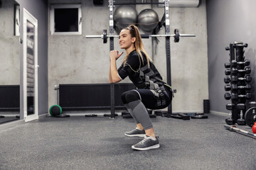A beautiful woman in an EMS suit does squats in a gym. - obrazy, fototapety, plakaty
