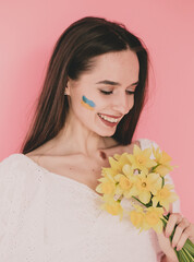 
Woman of model appearance with the flag of Ukraine on her face holds yellow flowers in her hands and smiles on a pink background
