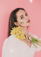 Woman of model appearance with the flag of Ukraine on her face holds in her hands yellow flowers on a pink background