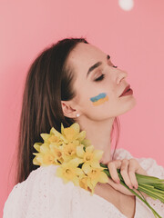 Woman of model appearance with the flag of Ukraine on her face holds in her hands yellow flowers on a pink background