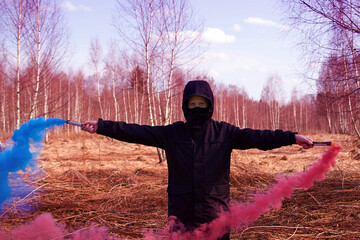 A preteen in a black jacket and a mask holds two colored smoke bombs