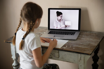 Cute European girl writes at table with laptop, video chat with teacher, distance learning and tutor, self-learning by video lesson