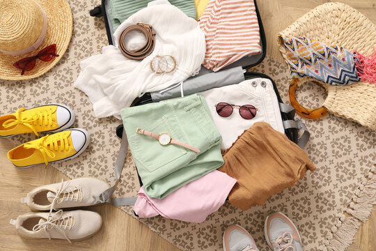 Open suitcase full of clothes and summer accessories near shoes on floor, top view