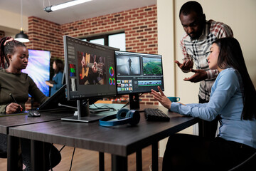 Footage editing specialist using multi monitor workstation to improve frames quality. Professional videographer sitting at desk while coworker helping her with film visual improvement.