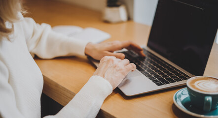 Mature adult woman hands, sitting in cafe with coffee mug and working online on laptop. Businesswoman typing on notebook computer in coworking space in roasters coffee shop, close up