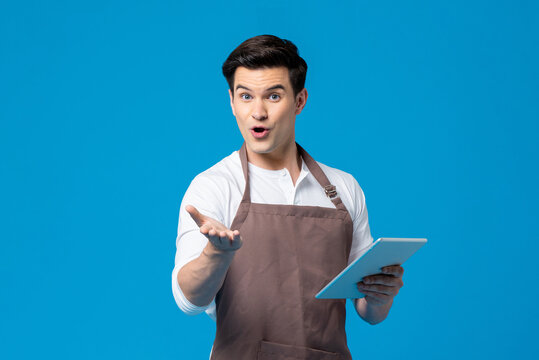 Serviced minded Caucasian male waiter with apron holding tablet and taking order on blue background in light studio