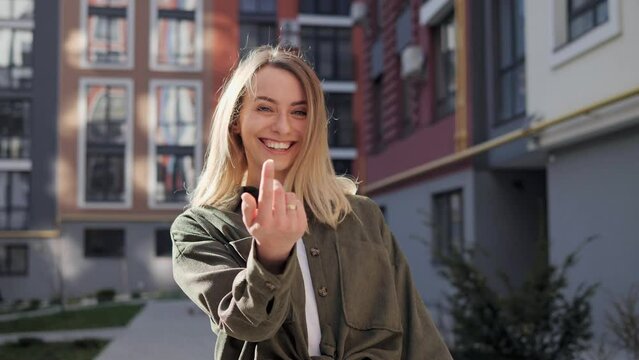 Gorgeous girl waving hand to come with me on city street. Come here, follow me. Beautiful young blond woman makes a beckoning gesture with one finger, inviting to approach, looks playfully flirting.