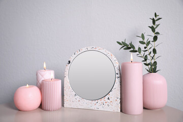 Small mirror, burning candles and vase on table near light wall