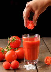 A glass cup with tomato juice, next to it lies a branch of tomatoes. On a wooden table there is a glass of tomato juice and tomatoes lie on a black background.Hand holding a tomato over a glass of jui