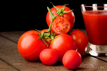 A glass cup with tomato juice, next to it lies a branch of tomatoes. On a wooden table there is a glass of tomato juice and tomatoes lie on a black background.