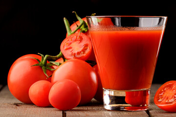 A glass cup with tomato juice, next to it lies a branch of tomatoes. On a wooden table there is a glass of tomato juice and tomatoes lie on a black background.