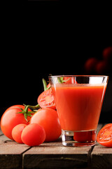 A glass cup with tomato juice, next to it lies a branch of tomatoes. On a wooden table there is a glass of tomato juice and tomatoes lie on a black background.
