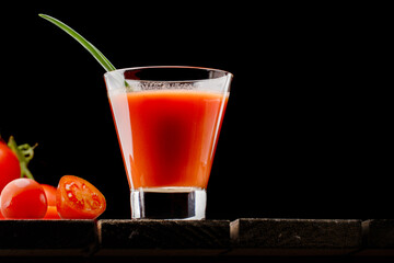 A glass cup with tomato juice, next to it lies a branch of tomatoes. On a wooden table there is a glass of tomato juice and tomatoes lie on a black background.