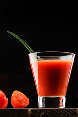 A glass cup with tomato juice, next to it lies a branch of tomatoes. On a wooden table there is a...