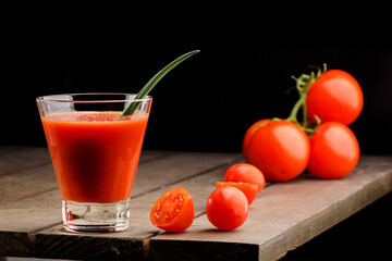 A glass cup with tomato juice, next to it lies a branch of tomatoes. On a wooden table there is a...