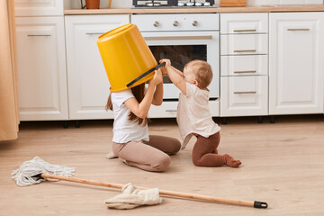 Indoor shot of two little cute girls playing in kitchen on floor, elder girl putting yellow bucket...
