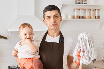 Portrait of father and infant baby daughter with floor mops in kitchen, man wearing brown apron...