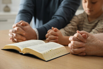 Boy and his godparents praying together at wooden table indoors, closeup