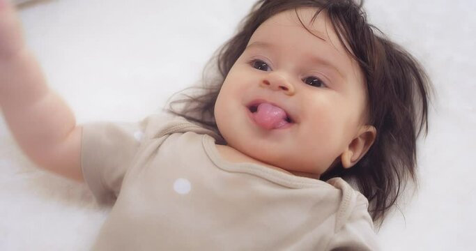 Close-up.Happy baby girl is lying on her back on a white bed, dressed in a beige bodysuit with white peas, smiling from her mother's tickling, playing with her mother, showing her tongue 