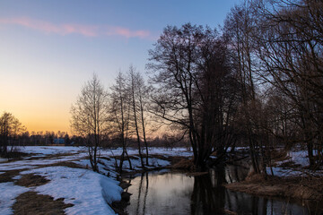 Dramatic sunset view over the river in winter; colorful sky and soft evening light; trees are reflected in the water