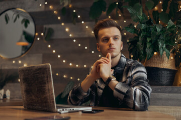 Pensive young man sitting at table with laptop and thinking about project. Male freelancer...