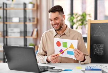 distance education, primary school and remote job concept - happy smiling male teacher with laptop and picture of geometric shapes in different colors having online class at home office - obrazy, fototapety, plakaty