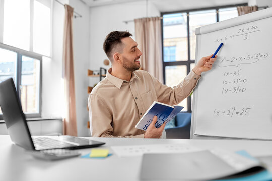 distant education, school and remote job concept - happy smiling male math teacher with laptop compute and book showing equation on flip chart having online class or video call at home office