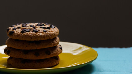 Pile of chocolate chip cookies on a yellow plate and on a blue tablecloth and dark background