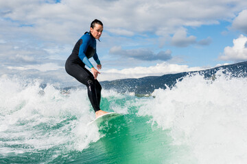 Athletic woman in wetsuit riding on endless waves behind a boat on sunny day. Female learning...