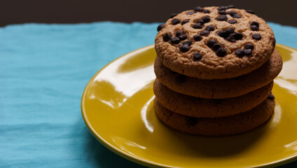 Pile of chocolate chip cookies on a yellow plate and on a blue tablecloth and dark background