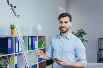 Portrait of a successful business founder, a man looks at the camera and smiles at a white board for notes, holding a tablet, describes a business strategy
