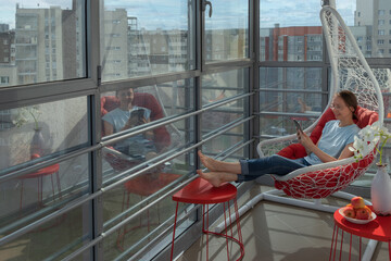 Barefoot pretty woman in blue t-shirt and jeans sits on hanging wicker white chair with red cushion...