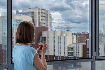Fototapeta premium Back view of caucasian woman in blue t-shirt stands at panoramic window with cup of hot tea and looks at city landscape. Concept of rest, vacation, opportunities, success, sightseeing and future.