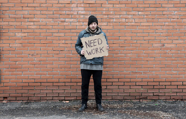 Homeless holds piece of cardboard with inscription Need Work. Refugee is at brick wall looking for...