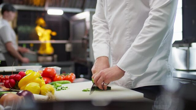 Cropped shot of hands of unrecognizable restaurant chef cutting fresh cucumber on kitchen table as his colleague flambeing food in background