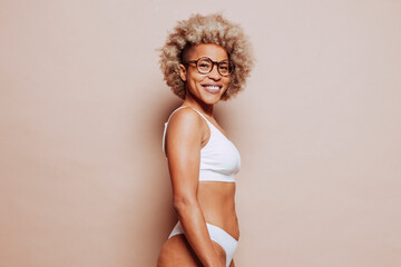 Side view portrait of young happy smiling afro latin american woman in white underwear and eyeglasses over beige background at studio. Real people, real body, natural beauty.
